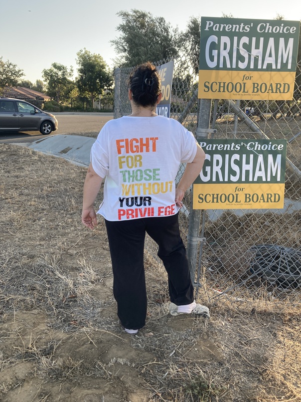 Jennifer Grisham, wearing a shirt that reads "Fight for those without your privilege," standing beside Grisham for School Board yard signs